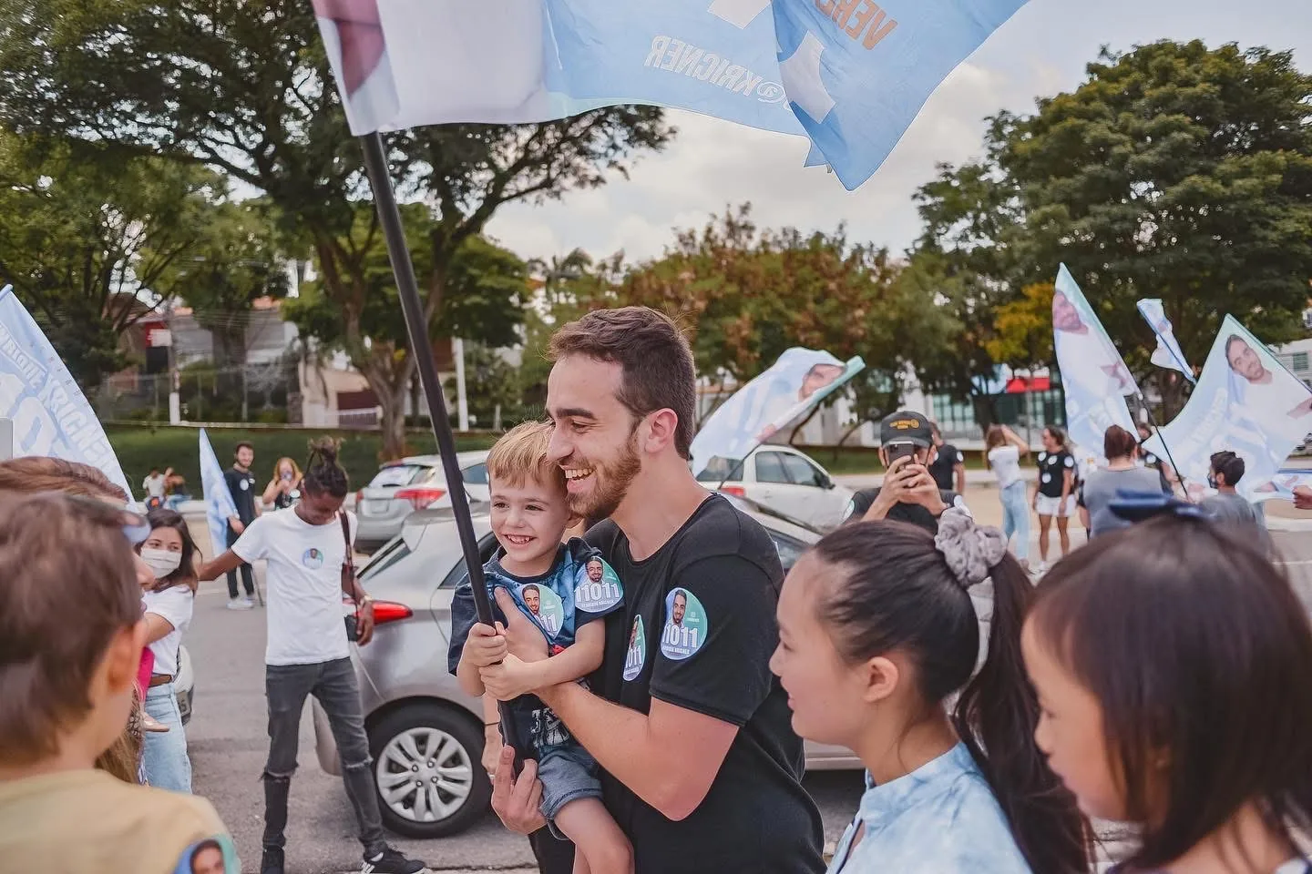 Henrique Krigner sorri segurando uma criança no colo, cercado de apoiadores com bandeiras da campanha de 2020